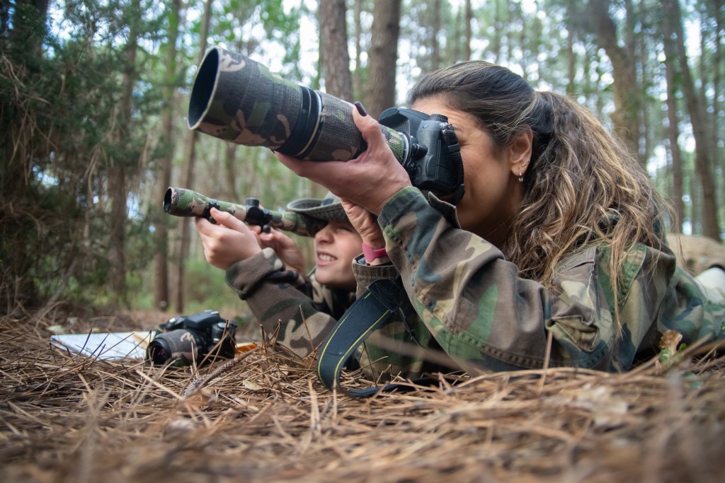 alt="Absorbed mother and son taking pictures in the forest. Family with modern cameras lying on the ground, using camera and binoculars. Parenting, family, leisure concept."